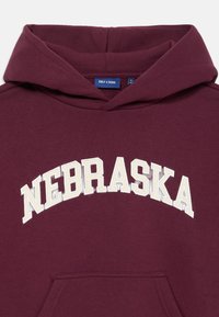 Bordová mikina s kapucňou a veľkým oblúkovým logom "NEBRASKA" v smotanovej farbe. Obsahuje šnúrku na kapucni a prednú vrecko. Mäkká textúra látky.