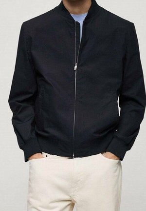 Uomo che indossa una giacca bomber nera con zip sopra una camicia azzurra e pantaloni bianchi, con le mani in tasca, in piedi contro uno sfondo semplice.