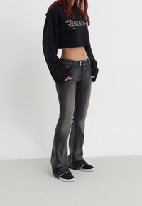 Persona que lleva jeans acampanados de color negro desgastado, una camiseta de manga larga negra corta con texto y zapatillas negras sin cordones, frente a un fondo blanco liso.