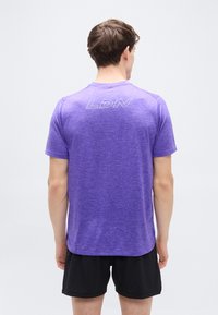 Μωβ αθλητική μπλούζα με κοντά μανίκια και στρογγυλή λαιμόκοψη, με αντανακλαστικό τύπωμα "LDN" στο πάνω μέρος της πλάτης. Φορεμένη με μαύρο σορτς.