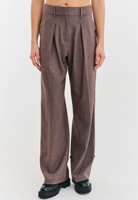 Osoba oblečená v hnedých nohaviciach s vysokým pásom a voľným strihom s záhybmi, v kombinácii s čiernymi topánkami s hrubou podrážkou, stojaca na jednofarebnom pozadí.