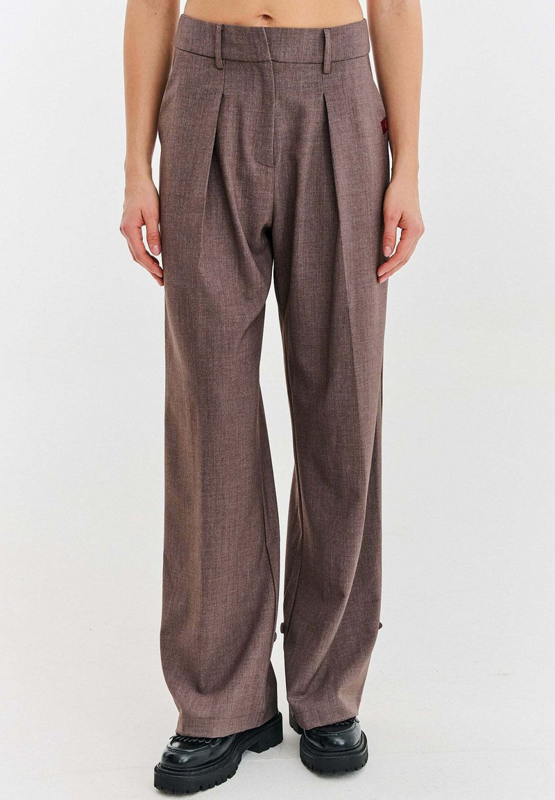 Osoba oblečená v hnedých nohaviciach s vysokým pásom a voľným strihom s záhybmi, v kombinácii s čiernymi topánkami s hrubou podrážkou, stojaca na jednofarebnom pozadí.