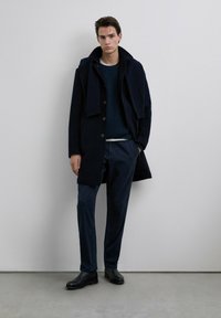 Navy overcoat with buttons, layered over a dark sweater, paired with matching trousers and black leather boots. Smooth texture and fitted design.
