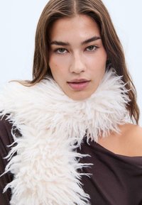 Sciarpa bianca e morbida, con un design testurizzato, avvolta attorno al collo, abbinata a un indumento marrone scollato. Aspetto morbido e voluminoso.