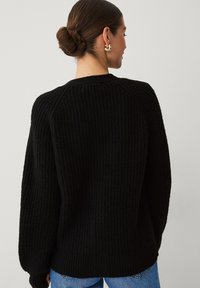 Zwarte gebreide trui met een geribbelde structuur, ronde hals en drop-shoulder mouwen. De achterkant is iets langer en wordt gedragen met een blauwe jeans.
