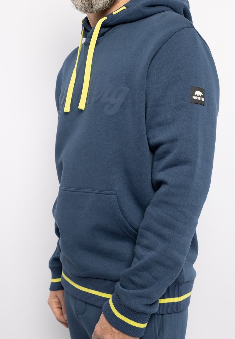 Homme portant un sweat à capuche bleu avec des cordons jaunes et des bandes jaunes aux poignets et à l'ourlet, affichant un patch de la marque Forsberg sur la manche.