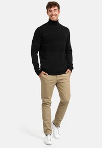 Maglione nero a collo alto con strisce testurizzate, abbinato a pantaloni chinos beige e sneakers bianche, che mostrano un design casual e aderente.