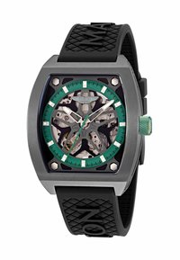 Reloj cuadrado de acero inoxidable con bisel verde, esfera esqueletizada que muestra engranajes, marcadores blancos, correa negra texturizada y corona verde.