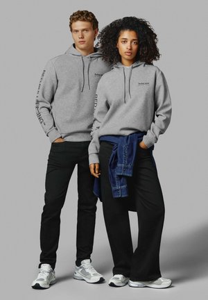Des sweat-shirts gris avec cordons et logo, un pantalon noir et des baskets grises. Une personne porte une chemise en denim nouée autour de la taille.