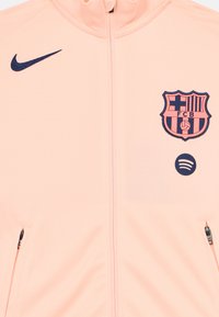 Nike jaka, kas izgatavota no mīksta persiku krāsas auduma, ir ar pilnu rāvējslēdzēju, sānu kabatām un izšūtu FC Barcelona logotipu ar ziliem akcentiem un Spotify emblemu.