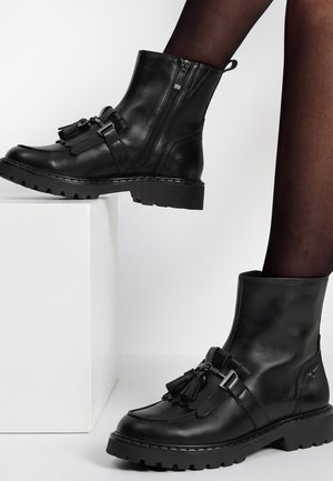 Bottines en cuir noir avec décorations en pompons et semelles épaisses portées avec des collants noirs transparents, un pied reposant sur un bloc blanc.