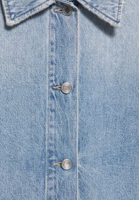 Light blue denim jacket front with metal snap buttons, visible stitching, and a collar with slightly frayed edges.