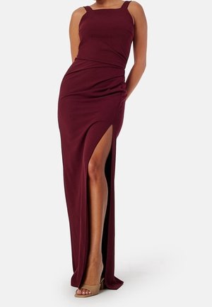 Robe de soirée bordeaux sans manches avec une fente haute sur la jambe gauche, portée avec des talons ouverts couleur nude sur une femme debout.