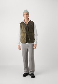 Olīvu zaļa, viegla veste ar vairākiem rāvējslēdzējiem kabatām un priekšējo rāvējslēdzēju, uzvilkta virs baltas garām piedurknēm krekla un pelēkiem biksēm.