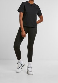 Persoon in een zwart T-shirt met korte mouwen, zwarte leggings, witte sokken en witte sneakers staat tegen een effen grijze achtergrond.