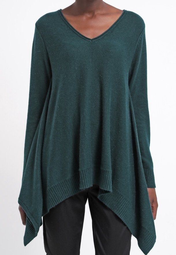 Pull en maille vert foncé à manches longues avec col en V et ourlet asymétrique en forme de mouchoir porté sur un pantalon noir.