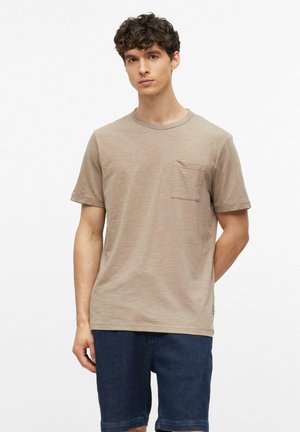 Jeune homme aux cheveux bouclés portant un t-shirt beige à manches courtes avec une poche poitrine et un short bleu foncé, debout devant un fond uni.