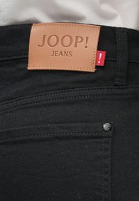 Μαύρο τζιν με δερμάτινη ετικέτα που φέρει ανάγλυφη την επιγραφή "JOOP! JEANS" και κόκκινη ετικέτα διακοσμητική. Περιλαμβάνει κανονικές τσέπες και μεταλλικά εξαρτήματα.