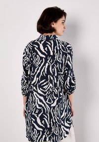 Una donna con capelli corti è di spalle, indossa una lunga camicia a fantasia con righe blu navy e bianche, su uno sfondo bianco semplice.