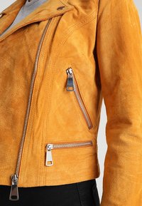 Gul mockaläder motorcykeljacka med silverdragkedjor, cropped design och sidofickor med dragkedja. Mjuk textur och synliga sömnadsdetaljer.