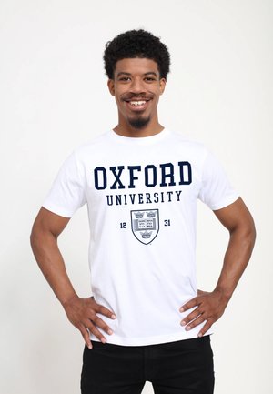 Smilende mand med kort krøllet hår står med hænderne på hofterne iført en hvid Oxford University t-shirt og sorte bukser foran en ensfarvet baggrund.