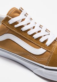 Bruine canvas sneaker met witte veters, een witte leren zijstreep en een contrasterende rubberen zool. Heeft een lichtbruine stiksels en platte veters.
