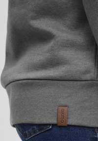 Primer plano de una persona que lleva una sudadera gris con una etiqueta marrón "oxmo" y jeans de mezclilla azules con costuras visibles.