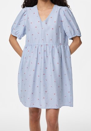 Donna che indossa un vestito azzurro a righe con piccole stampe floreali rosse, maniche corte a sbuffo e scollatura a V, in piedi con le mani dietro la schiena.