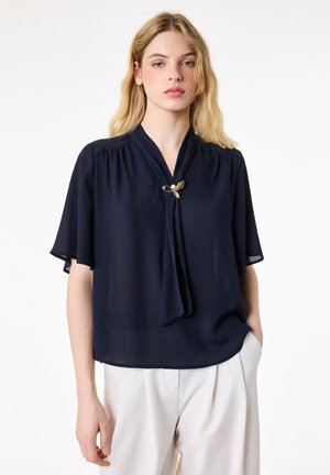 Femme blonde portant un chemisier à manches courtes bleu marine avec une broche en or et perles, associée à un pantalon blanc, debout devant un fond blanc.