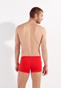 Un maillot de bain rouge ajusté, fabriqué à partir d'un matériau lisse. Dispose d'une étiquette bleue à l'arrière, au design minimaliste sans motifs ni accents.