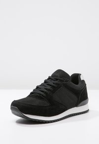 Chaussure de sport noire en mélange de matériaux en maille et en daim, avec un design texturé, semelle en caoutchouc et accents blancs sur le talon et le midge.