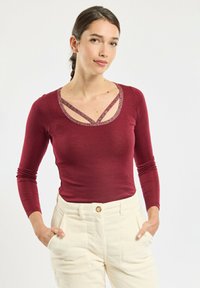 Burgundy long-sleeve top with a scoop neckline featuring crisscrossed glitter accents; smooth fabric, form-fitting design.