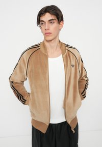 Sametová bomber bunda v béžové barvě s černými pruhy na rukávech, ribbonem na zápěstích a spodním lemu. Obsahuje decentní logo na hrudi a přední zipové zapínání.