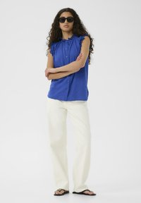 Kék pánt nélküli blúz fodros vállakkal, gombokkal az elején. Fehér, bő szárú nadrággal és fekete papuccsal párosítva.