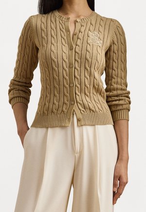 Kobieta w beżowym swetrze na guziki z wzorem warkocza i wyszywanym logo, w połączeniu z kremowymi, szerokimi spodniami z wysokim stanem.