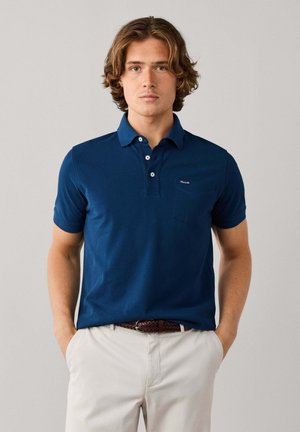 Joven con cabello ondulado vestido con un polo azul marino y pantalones beige claros, con las manos en los bolsillos, sobre un fondo liso.
