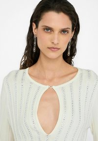 Maglione chiaro con scollo a V profondo e design a occhio di serratura, decorato con paillettes argentate verticali e un dettaglio con chiusura metallica.