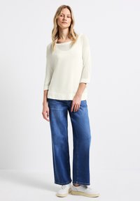 Maglione chiaro con scollatura ampia e maniche a tre quarti; abbinato a jeans blu larghi e sneaker bianche.