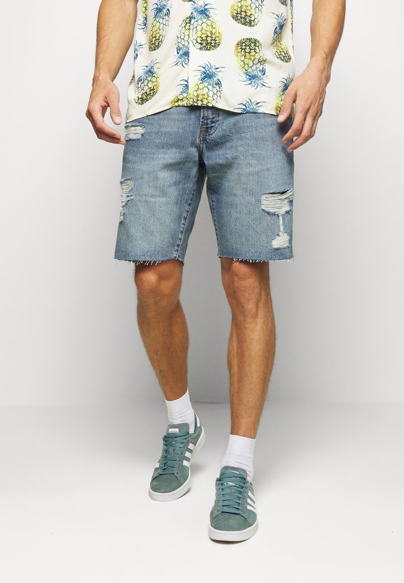 Homme portant un short en jean usé, des chaussettes blanches, des baskets vertes et une chemise avec des motifs d'ananas jaunes sur fond blanc.