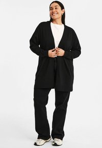Svart cardigan med öppen fram, långa ärmar och en avslappnad passform, bärs över en vit t-shirt och svarta byxor, tillsammans med sneakers.
