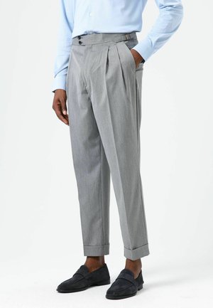 Pantalon gris plissé à taille haute, équipé de doubles boutons et de passants de ceinture, terminé par des ourlets retroussés. Porté avec des mocassins foncés.