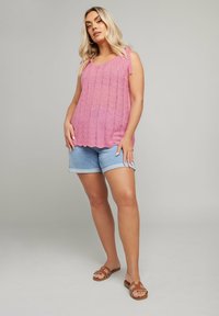 Rosa, stickad linne med öppet stickat mönster och knytband på axlarna; matchat med ljusa jeansshorts och bruna sandaler.