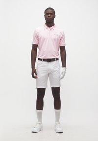 Homme portant un polo rose clair, un short blanc, un gant de golf blanc à la main gauche, des chaussettes blanches et des baskets blanches, debout devant un fond blanc.