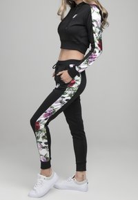 Femme en hoodie noir cropped et joggers assortis avec des bandes florales sur les côtés, portant des baskets blanches, se tenant de profil avec les mains dans les poches.