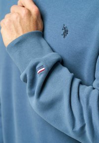 Kék pulóver bordás mandzsettával, a mellkasán egy kis hímzett logóval és a hosszú ujján egy háromszínű díszítő folttal.