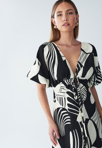 Schwarz-weißes Kleid mit tiefem V-Ausschnitt, kurzen Ärmeln und einer Schnürung in der Taille, versehen mit abstrakten Blattmustern und einem strukturierten Stoff.