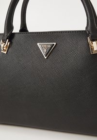 Sac à main en cuir noir texturé avec deux poignées et un logo triangulaire doré Guess USA au centre devant.