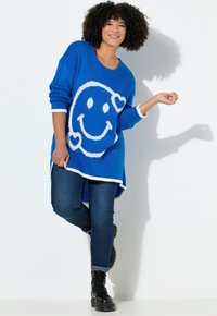 Blauwe gebreide trui met een groot wit smiley gezicht ontwerp, voorzien van een ronde hals, lange mouwen en geribbelde witte accenten. Gecombineerd met donkere jeans en zwarte veterschoenen.