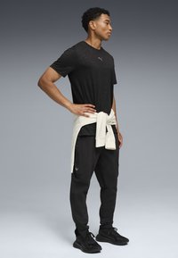 Sort atletisk t-shirt med subtilt mønster, beige sweatshirt bundet om taljen, sorte tapered bukser og sorte sneakers.