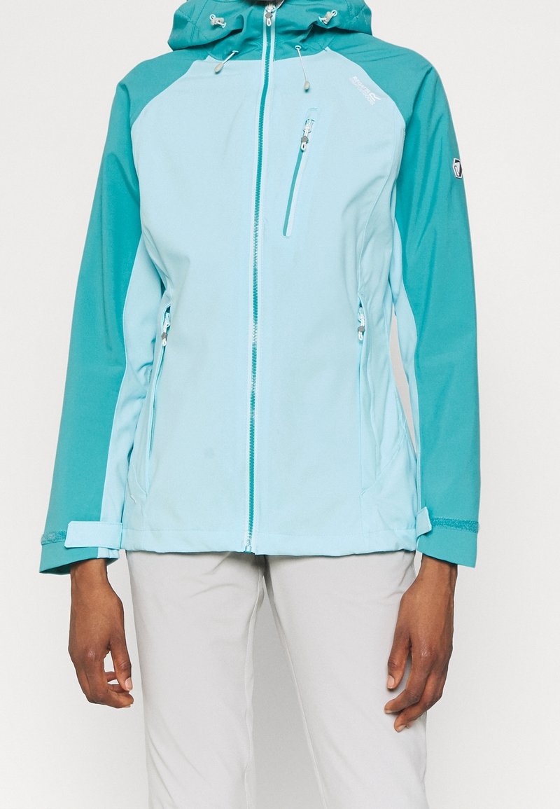 Veste à capuche bleu clair et bleu sarcelle avec fermeture éclair devant et poches zippées, portée par une personne avec un pantalon clair, montrée du cou aux hanches.